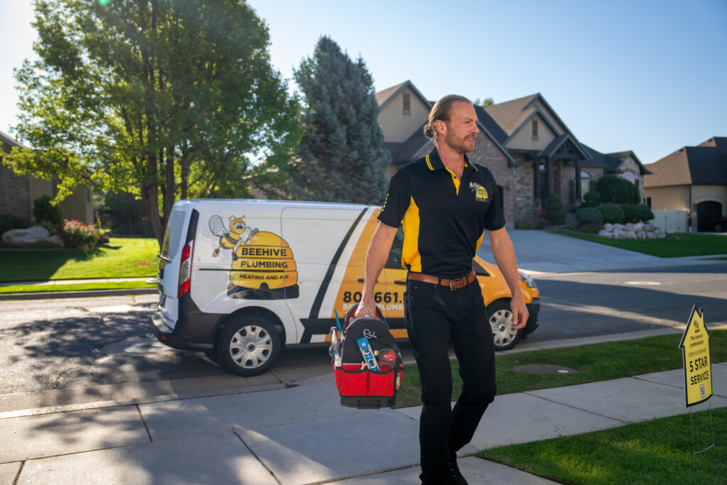 man in beehive plumbing uniform carrying red tool bag walking up a driveway