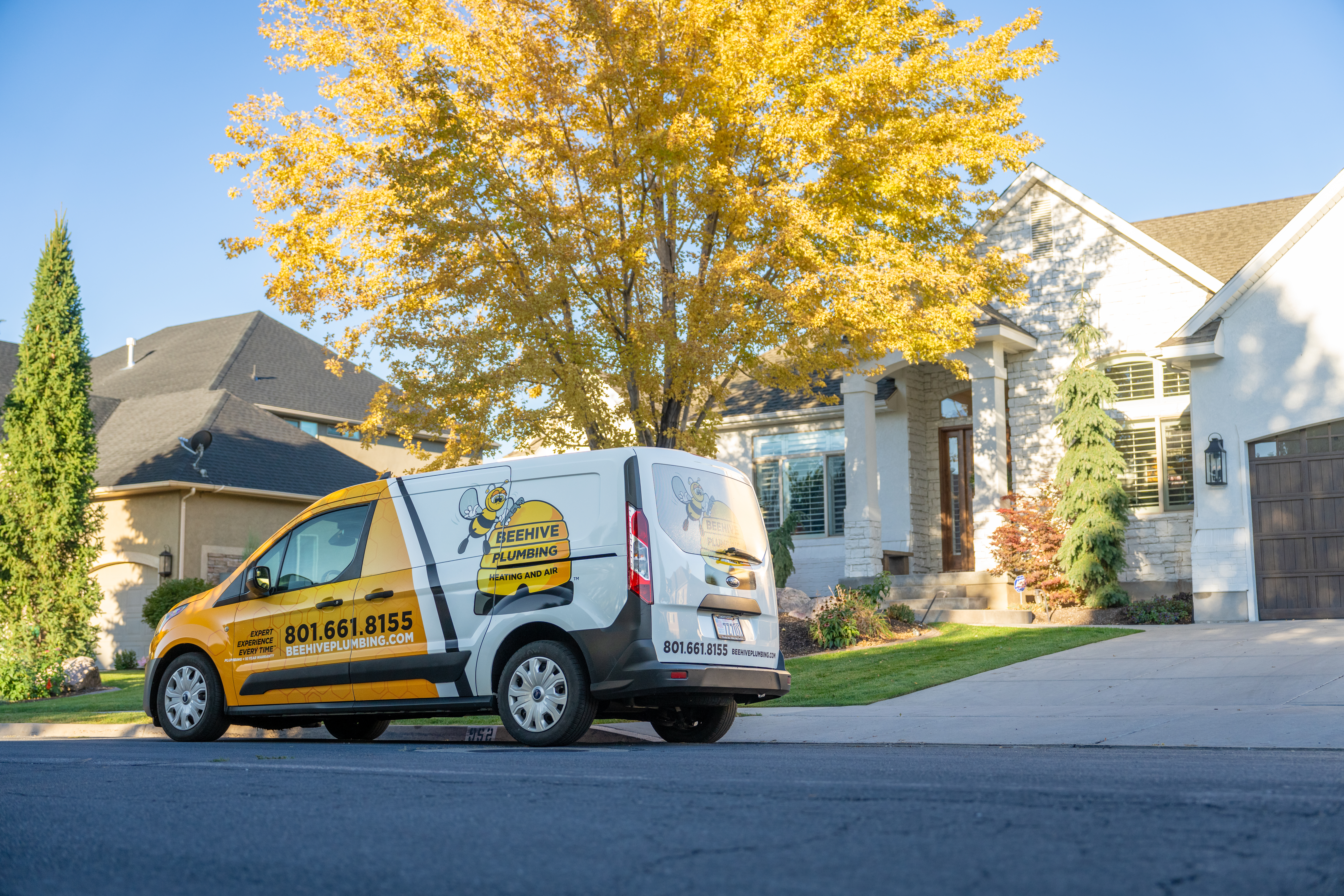 A Beehive Plumbing service van is parked in front of a beautiful suburban home with a well-maintained lawn and landscaping. The van is white and yellow, featuring the company's logo, contact number, and website. A large tree with golden autumn leaves stands in the yard, and the house has a stone and stucco exterior with large windows and a wooden front door. The setting is a quiet residential neighborhood on a sunny day.