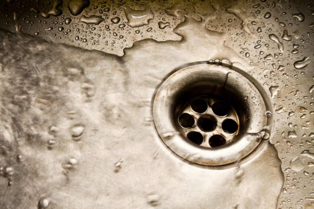 A close-up of a stainless steel sink drain with water droplets and small streams of water flowing around it. The surface of the sink appears slightly worn, with visible scratches and reflections. The lighting highlights the texture of the metal and the movement of the water.