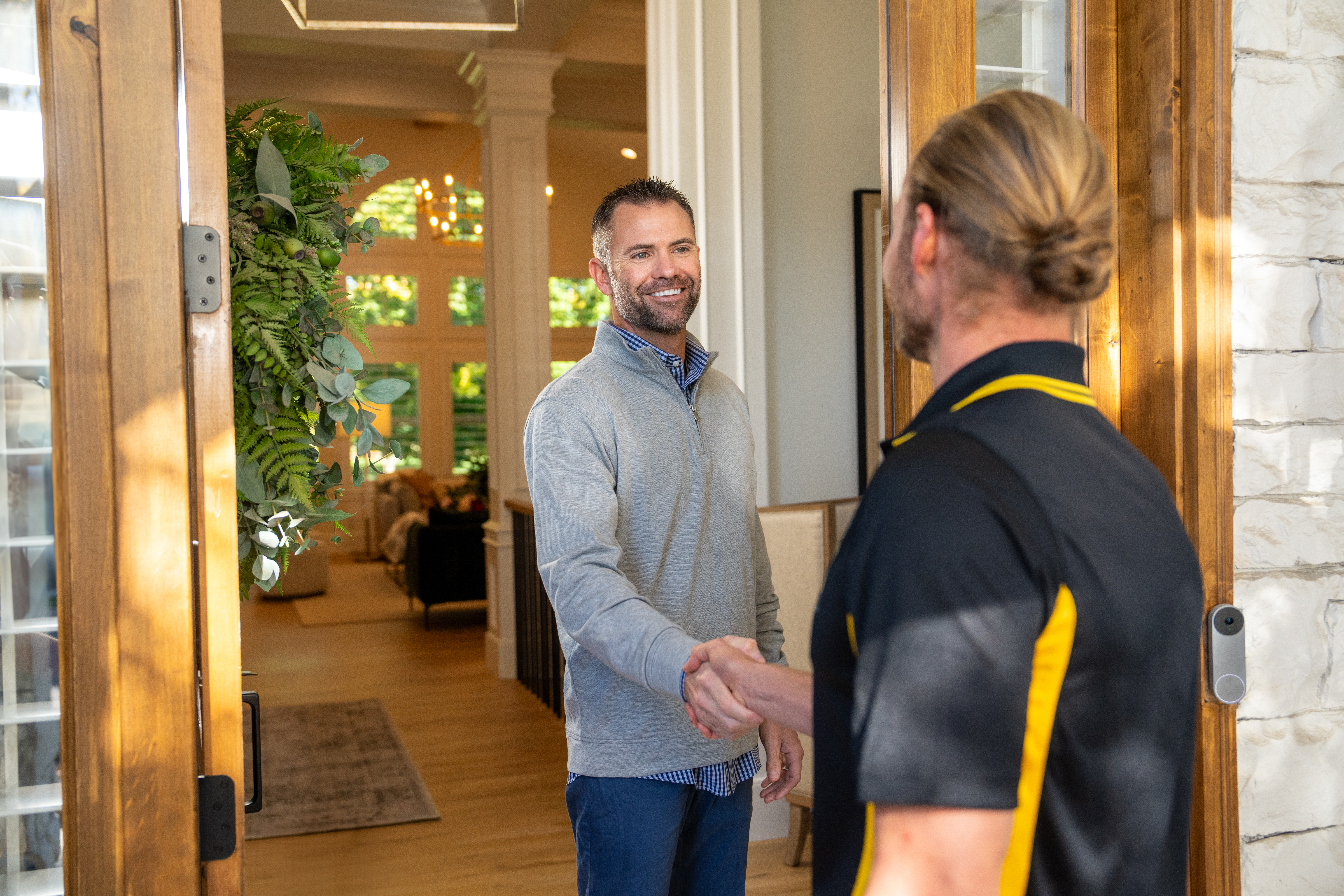 A smiling homeowner wearing a gray sweater shakes hands with a technician dressed in a black and yellow Beehive Plumbing uniform at the front door of a well-lit, elegant home. The entrance features wooden doors with glass panels and a decorative green wreath. The interior is warmly lit, showcasing a spacious living area with tall windows, chandeliers, and stylish furnishings. The interaction suggests a professional service visit, likely for plumbing or HVAC maintenance.