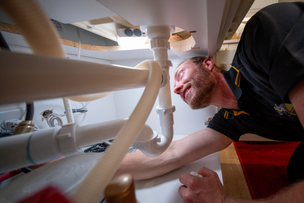 A plumber wearing a black and yellow Beehive Plumbing uniform is working under a sink, inspecting the white PVC pipes and connections. He has a focused expression as he reaches into the plumbing system, likely performing maintenance or repairs. The image is taken from a low angle, emphasizing the complexity of the pipes. The background shows part of a kitchen with a red rug on the floor.