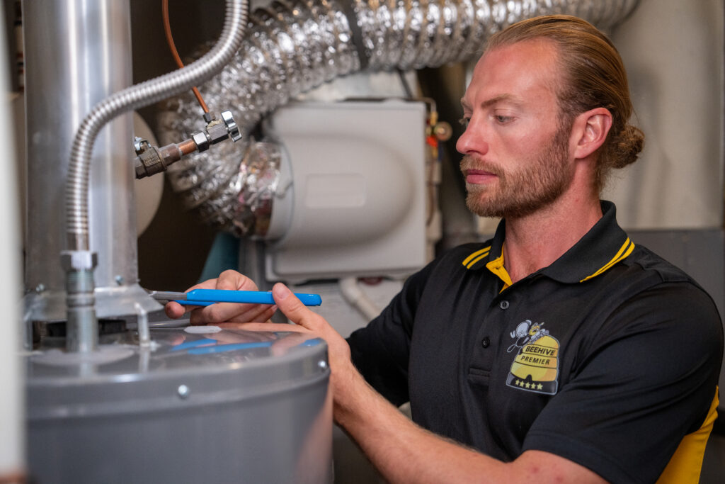 A technician with a beard and tied-back hair is working on a water heater in a utility room. He is wearing a black and yellow Beehive Premier polo shirt and using a wrench to make adjustments. The background includes ductwork, pipes, and HVAC equipment. He has a focused expression as he performs maintenance or repairs on the system.