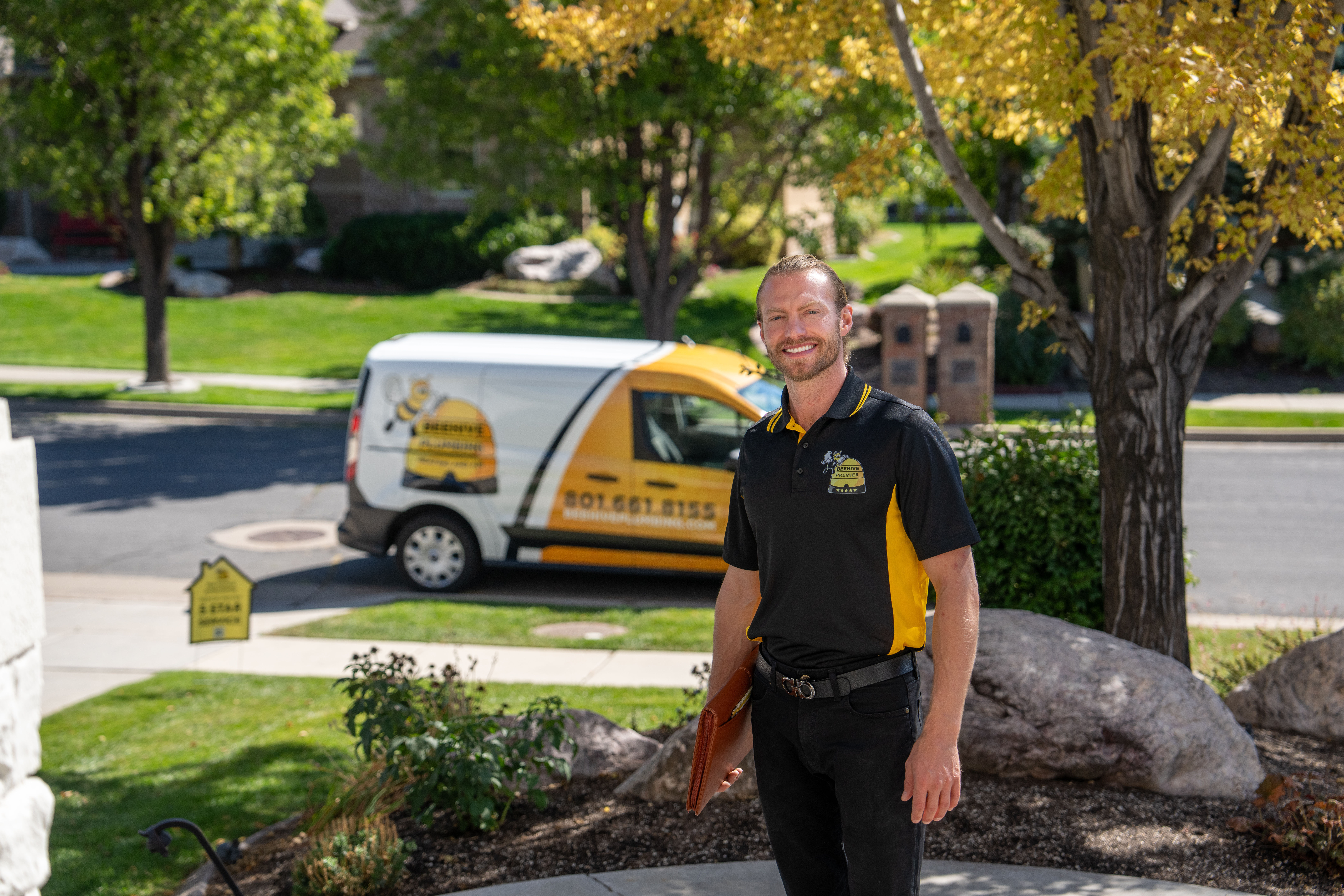 A smiling technician wearing a black and yellow Beehive Premier polo shirt stands in front of a home, holding a clipboard. Behind him, a company-branded service van with a bee-themed logo and contact information is parked on the street. The setting is a well-maintained suburban neighborhood with green lawns, trees, and landscaping. The technician appears professional and ready for a service appointment.