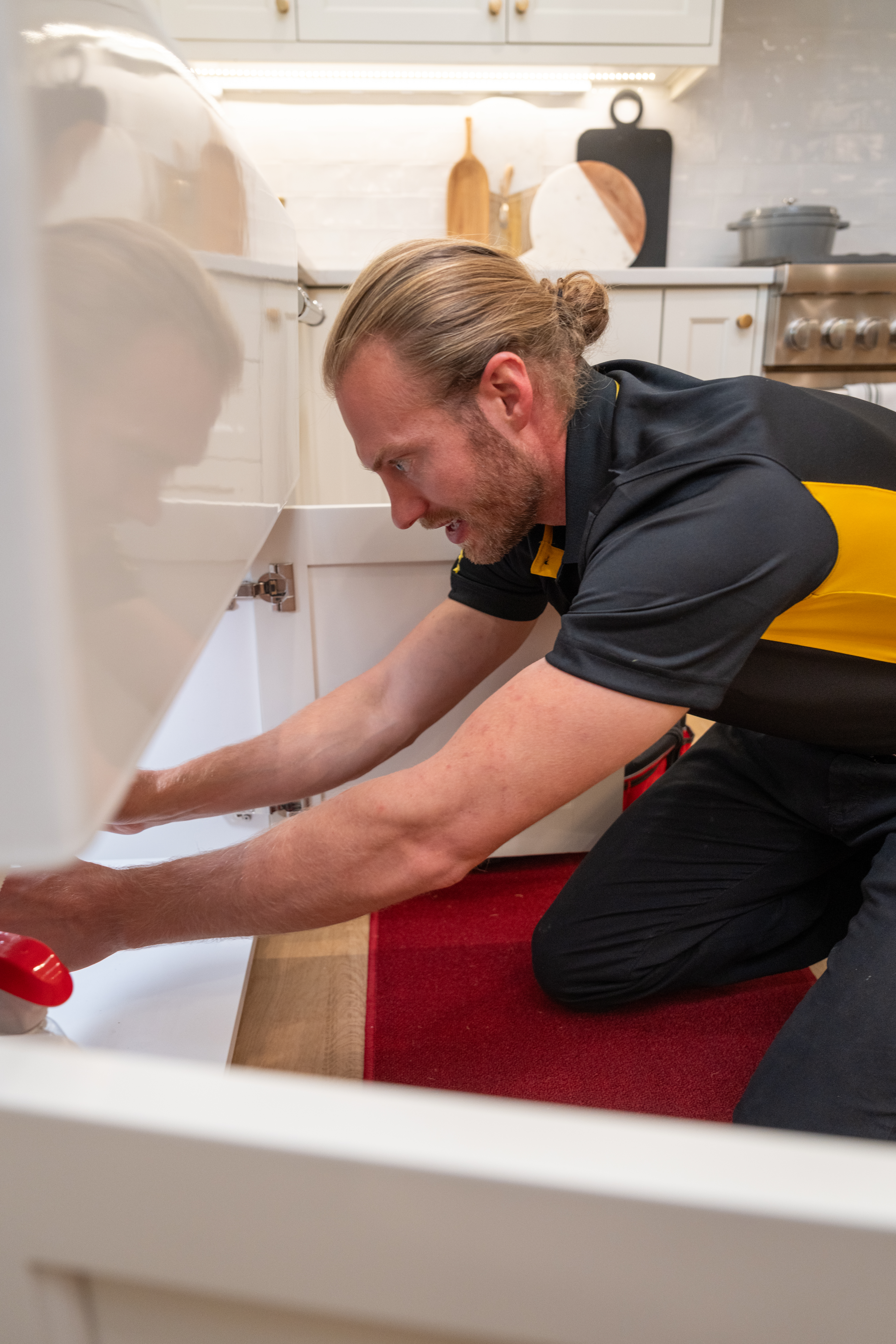 Image of a man working under a kitchen sink: A technician wearing a black and yellow uniform is crouched under a kitchen sink, making repairs. The modern kitchen features white cabinetry and wooden cutting boards on the counter.